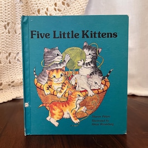 May include: A children's book titled "Five Little Kittens" with a teal cover. The cover illustration depicts five kittens in a woven basket, playing with a ball of yarn. The book is by Sharon Peters and illustrated by Amye Rosenberg.