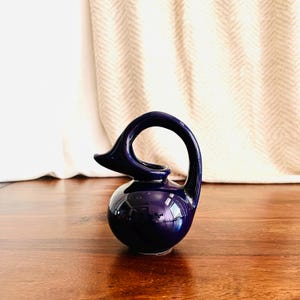 May include: A deep navy blue ceramic pitcher with a rounded, bulbous body and a singular, high-arched handle that curves gracefully from the top rim down to the base, creating a continuous, fluid silhouette with a pointed, beak-like spout.