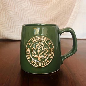 May include: A green ceramic mug with a gold emblem featuring a maple leaf and the words "Vermont Green Mountain State." The mug has a rounded shape and a curved handle. The mug is sitting on a wooden surface.