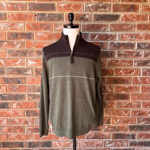 May include: A quarter-zip sweater featuring a dark brown upper chest and collar section, transitioning into a muted olive green lower body. A thin, horizontal cream-colored line separates the two colors across the chest, with long sleeves.