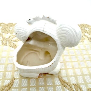 May include: White ceramic planter shaped like a stylized animal head. The planter has a large opening and two rounded ear-like features. The exterior is textured, and the interior is smooth. Some areas show signs of wear.