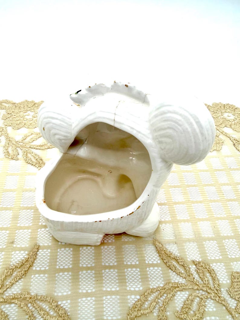 May include: White ceramic planter shaped like a stylized animal head. The planter has a large opening and two rounded ear-like features. The exterior is textured, and the interior is smooth. Some areas show signs of wear.