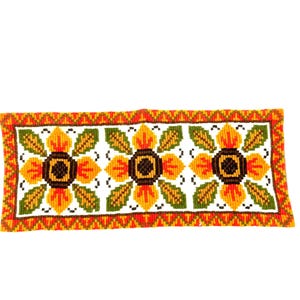 May include: Rectangular, hand-stitched textile featuring three large floral designs. Each flower has a brown center, yellow petals, and green leaves. The border is orange and green, with a white background. The textile measures approximately 12 inches by 6 inches.