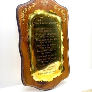 May include: A wooden wall plaque featuring a scalloped brass plate with the Ten Commandments engraved in black text. The wood has a dark brown finish with gold-painted leaf accents at the top. The brass plate has a reflective, wavy edge.