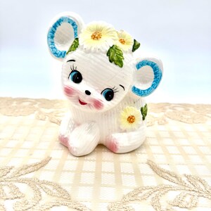 May include: A white ceramic mouse figurine with blue-trimmed ears and blue eyes. The mouse has a floral crown of yellow daisies and green leaves. Pink blush accents the cheeks and feet. The figurine is sitting on a cream-colored lace tablecloth.