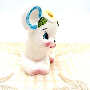 May include: A white ceramic mouse figurine with blue ears and a yellow flower. The mouse has pink cheeks and feet, and black and blue eyes. This decorative figurine is a charming collectable.