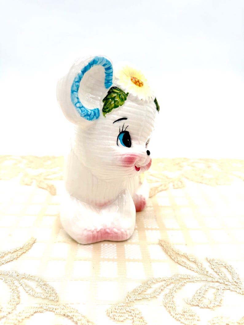 May include: A white ceramic mouse figurine with blue ears and a yellow flower. The mouse has pink cheeks and feet, and black and blue eyes. This decorative figurine is a charming collectable.