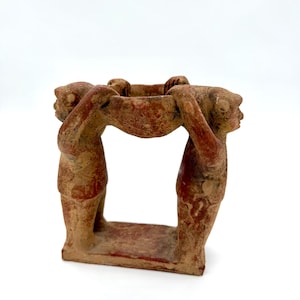May include: A terracotta sculpture featuring two figures supporting a bowl-shaped structure. The figures are depicted in a stylized manner, with visible details of their form. The sculpture has a reddish-brown hue and stands on a rectangular base.