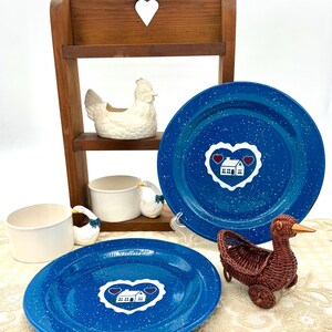May include: A collection of country-themed decor. Includes two blue enamel plates with white speckles and a heart-shaped house design, a wooden shelf, ceramic chicken and goose figurines, a white bowl, and a woven duck basket.