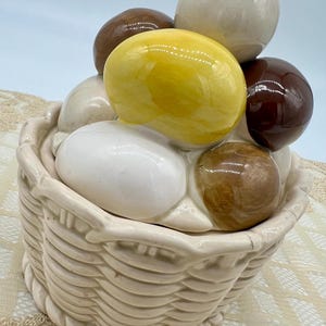 May include: A ceramic basket-shaped container with a lid, filled with glossy, egg-shaped ornaments in shades of white, brown, and yellow. The basket has a woven texture and a cream color.