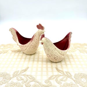 Peut inclure: Deux récipients en céramique en forme de poule. Les poules sont blanches avec des mouchetures rouges et des intérieurs rouges. Les récipients sont sur une surface crème à motifs.
