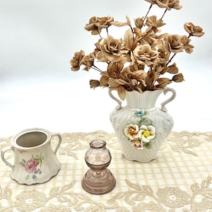 May include: A still life arrangement featuring a white ceramic vase with a floral design, filled with dried brown roses. A small pink glass bottle and a floral-decorated sugar bowl are also present.