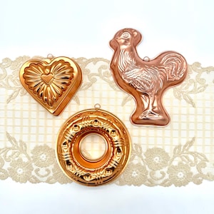 May include: Three copper-colored vintage molds: a heart with a central heart detail, a rooster, and a circular mold with a central hole. The molds are displayed on a cream-colored lace fabric.