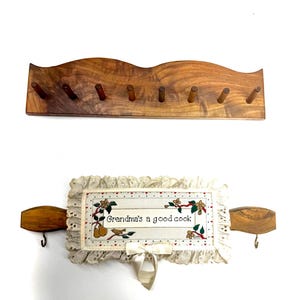 May include: A wooden wall rack with eight pegs and a rectangular fabric wall hanging featuring a ruffled lace border, a cream-colored center with the text "Grandma's a good cook," floral illustrations, and two wooden ends with metal hooks.