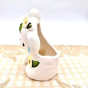 May include: A white ceramic bunny planter with a hollow interior. The bunny is decorated with yellow and green floral accents. The planter has a textured surface and is set against a neutral background.