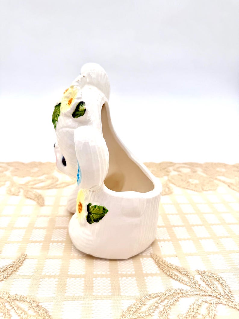 May include: A white ceramic bunny planter with a hollow interior. The bunny is decorated with yellow and green floral accents. The planter has a textured surface and is set against a neutral background.