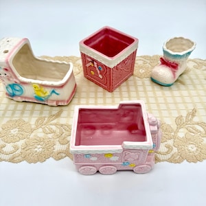 May include: Four ceramic baby-themed planters in a variety of shapes. The planters include a baby carriage, a square block, a baby bootie, and a train. The planters are primarily pink and white with decorative accents.