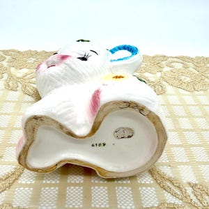 May include: A white ceramic bunny figurine with pink accents on the ears and cheeks, and blue on the inner ear. The bunny has a yellow flower on its side and the number 6189 on the base.