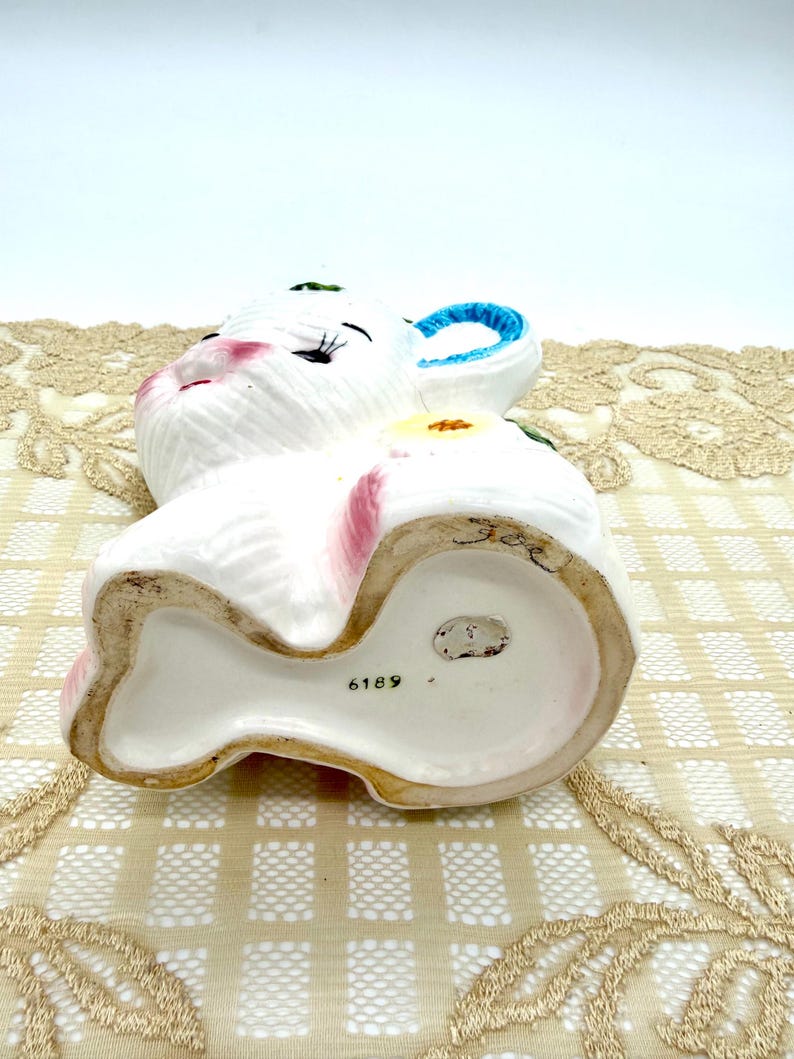 May include: A white ceramic bunny figurine with pink accents on the ears and cheeks, and blue on the inner ear. The bunny has a yellow flower on its side and the number 6189 on the base.