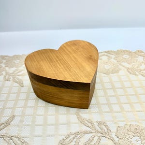 May include: A heart-shaped wooden box with a smooth, light brown finish and visible natural wood grain patterns. The box consists of a base and a matching lid, both crafted from wood, featuring a clean, simple design with rounded edges.