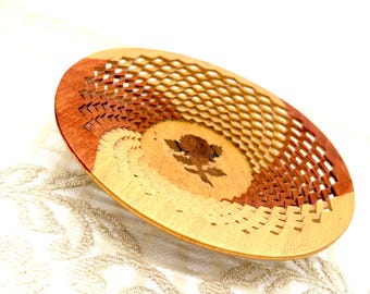 May include: A decorative wooden bowl with a unique design. The bowl features a light brown centre with a dark brown tree silhouette, surrounded by a geometric pattern and a reddish-brown rim. The bowl is on a white surface.