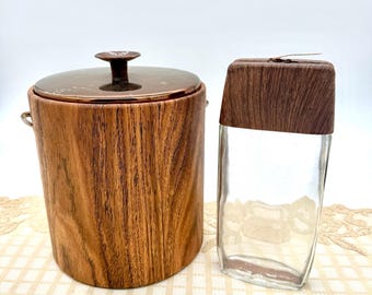 May include: A vintage ice bucket and a glass container with wooden accents. The ice bucket has a copper-coloured lid and handle, and a wood-grain finish. The glass container has a wooden lid. Both items are on a cream-coloured surface.