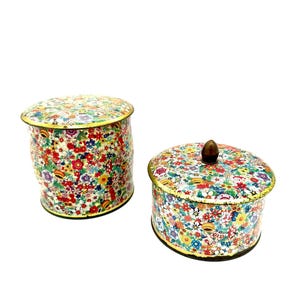 Peut inclure: Deux boîtes en fer blanc à motifs floraux. L'une est cylindrique, l'autre ovale, toutes deux avec des couvercles. Les couvercles et les côtés sont recouverts d'un motif floral coloré avec une bordure dorée. La boîte ovale a un bouton en bois sur le dessus.