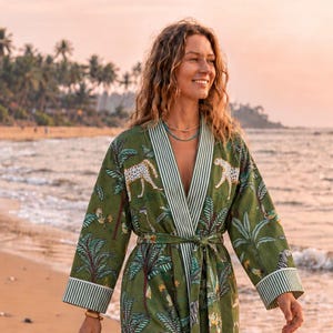May include: A long-sleeved olive green robe featuring a repeating print of white cheetahs and palm trees, with white and green striped trim on the collar and cuffs, a matching tie belt at the waist, and a tan fringed straw bag held in one hand.