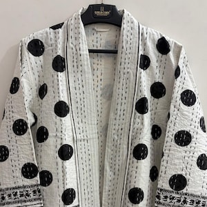 Puede incluir: Una chaqueta blanca abierta por delante con un patrón de lunares negros y detalles de costura vertical. La prenda incluye mangas anchas con un borde geométrico en blanco y negro en los puños y un ribete a juego en la abertura frontal.