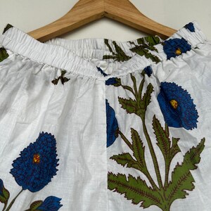 May include: White fabric pants with a blue floral pattern, featuring large blue flowers with green leaves and stems. The pants are hanging on a wooden hanger.