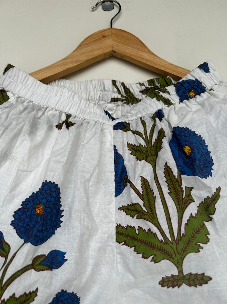 May include: White fabric pants with a blue floral pattern, featuring large blue flowers with green leaves and stems. The pants are hanging on a wooden hanger.