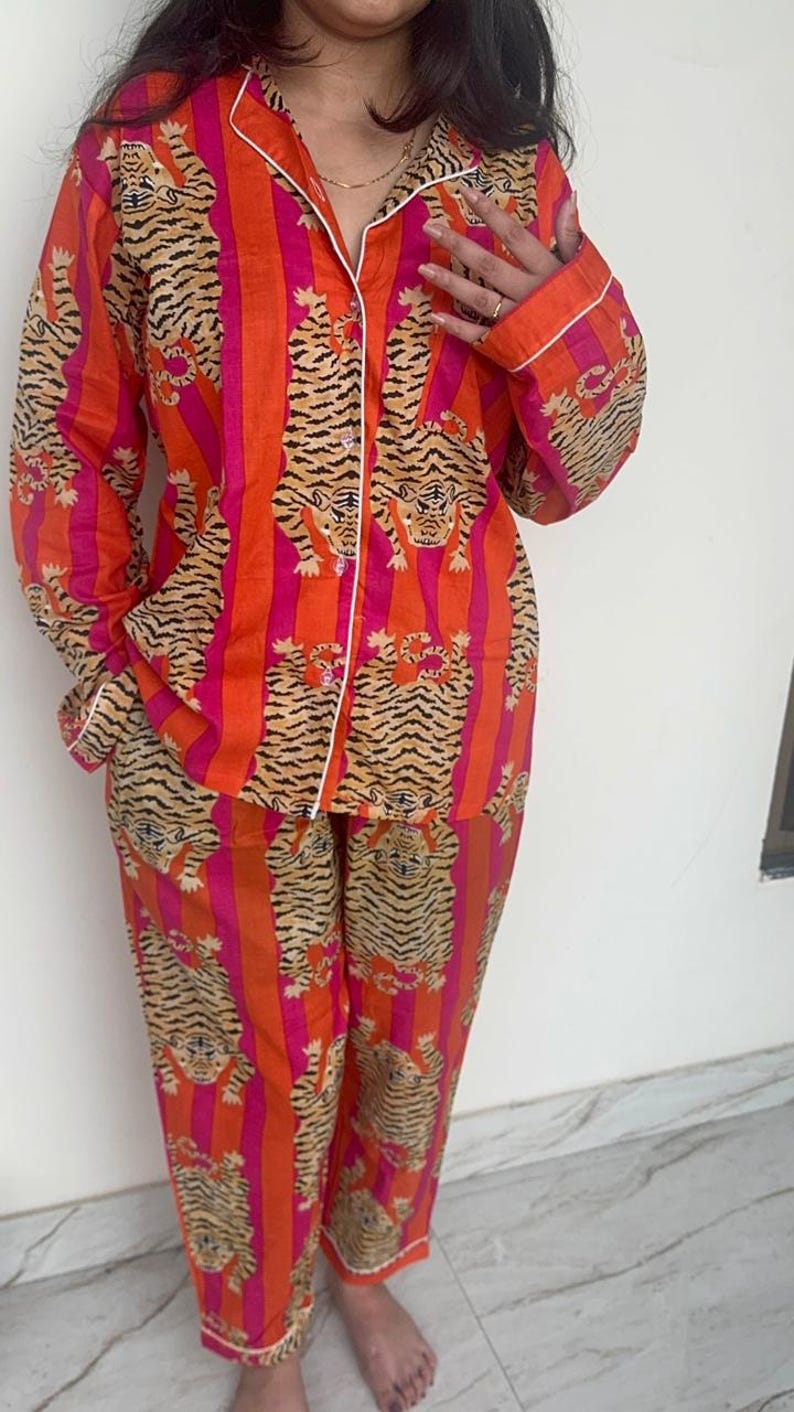 May include: A two-piece pajama set in a vibrant orange and pink striped pattern, featuring a repeating tiger print. The set includes a button-down shirt with white trim and matching trousers. The pajamas are made of a soft, comfortable material.