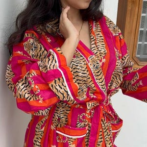 May include: A vibrant pink and orange striped robe with a tiger print design. The robe features a wrap-around style with a matching belt, white trim, and pockets. The long sleeves and knee-length cut make it a comfortable garment.