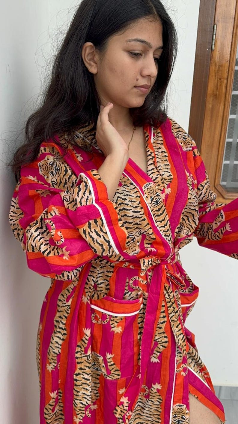 May include: A vibrant pink and orange striped robe with a tiger print design. The robe features a wrap-around style with a matching belt, white trim, and pockets. The long sleeves and knee-length cut make it a comfortable garment.