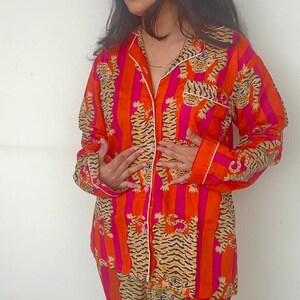 May include: An orange and pink striped pajama set featuring a tiger print. The long-sleeved top has a collared neckline and white trim. The matching trousers complete the set. The pajamas are made of a soft, lightweight fabric.