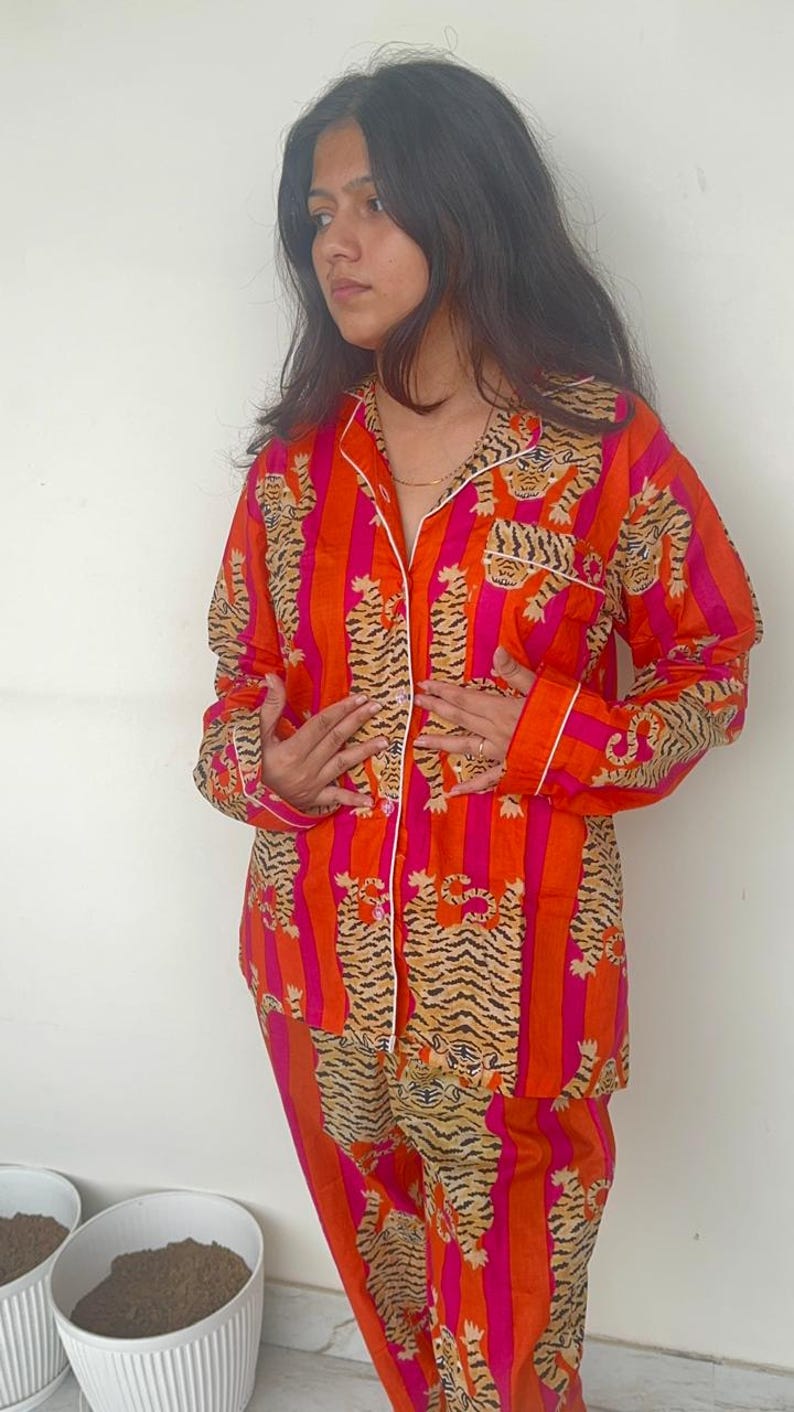 May include: An orange and pink striped pajama set featuring a tiger print. The long-sleeved top has a collared neckline and white trim. The matching trousers complete the set. The pajamas are made of a soft, lightweight fabric.