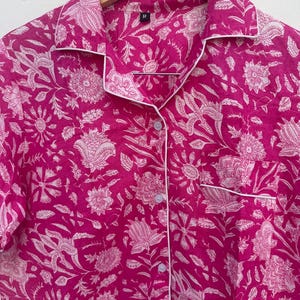 May include: A magenta pajama shirt with a white floral pattern and white trim. The shirt has a button-down front, a chest pocket, and a collar. The shirt is hanging on a wooden hanger.