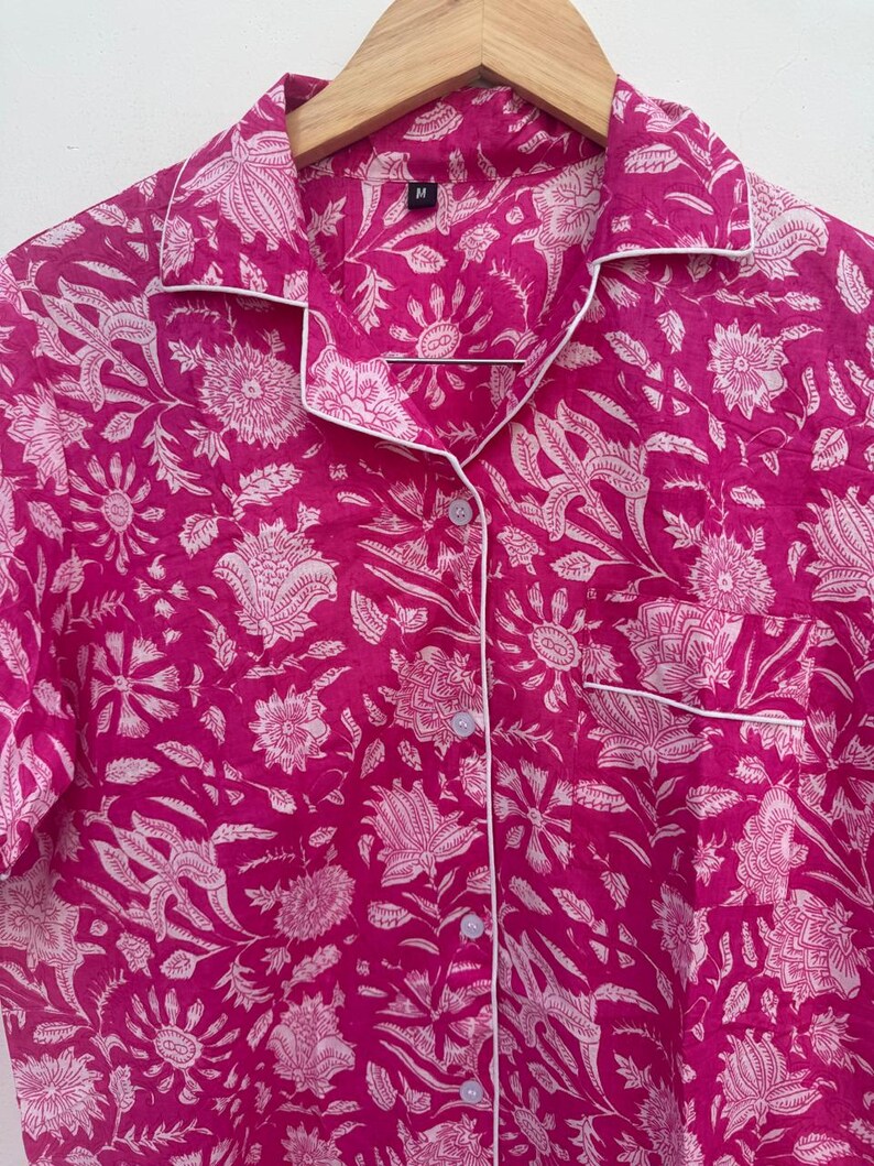 May include: A magenta pajama shirt with a white floral pattern and white trim. The shirt has a button-down front, a chest pocket, and a collar. The shirt is hanging on a wooden hanger.
