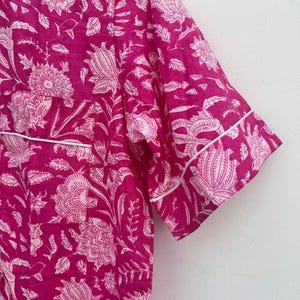 May include: A close-up of a magenta pajama top with a white floral pattern. The short-sleeved top has white piping along the edges. The fabric is a soft, lightweight material, perfect for sleepwear.