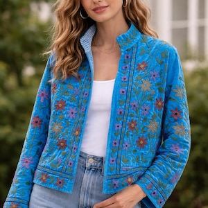 Puede incluir: Chaqueta azul claro con bordados florales en tonos rojos, morados y verdes. La chaqueta tiene cuello alzado y mangas largas. El forro es a rayas. La persona lleva un top blanco y vaqueros azules.