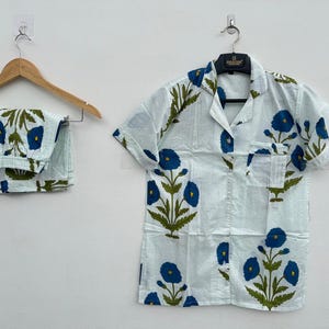 May include: A light blue pajama set with a floral pattern of blue flowers and green leaves. The set includes a short-sleeved button-down shirt and matching pants. The shirt and pants are hanging on a wooden hanger.