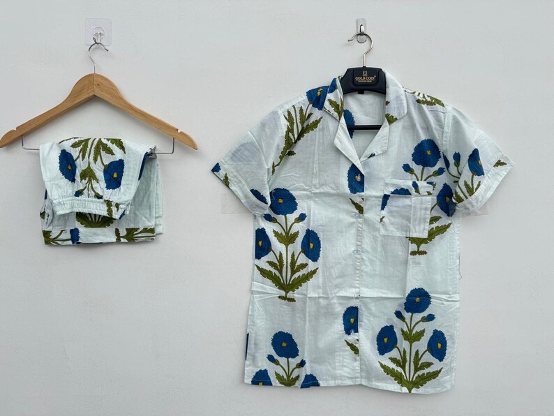 May include: A light blue pajama set with a floral pattern of blue flowers and green leaves. The set includes a short-sleeved button-down shirt and matching pants. The shirt and pants are hanging on a wooden hanger.