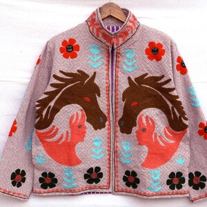 May include: A light purple jacket with a high collar and a zipper closure. The jacket features embroidered designs of brown horses, red flowers, and turquoise floral accents. The jacket is hanging on a wooden hanger.
