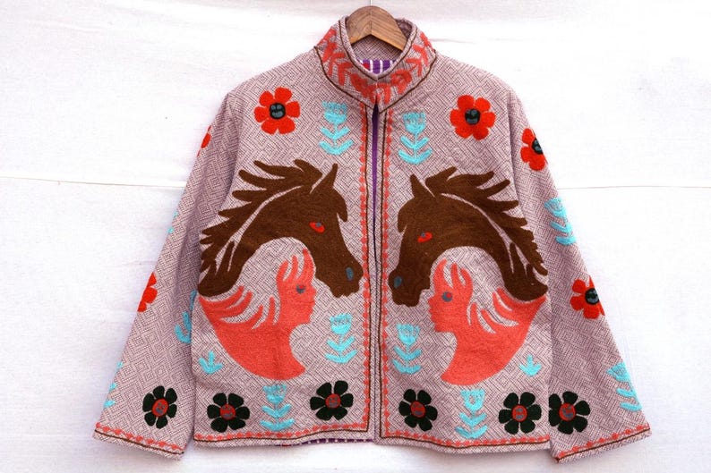 May include: A light purple jacket with a high collar and a zipper closure. The jacket features embroidered designs of brown horses, red flowers, and turquoise floral accents. The jacket is hanging on a wooden hanger.