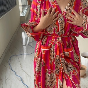 May include: A vibrant, long robe in shades of pink and orange, featuring a tiger stripe pattern. The robe has a wrap-around design with a matching belt, white trim, and pockets. The sleeves are long, and the robe is made of a lightweight fabric.