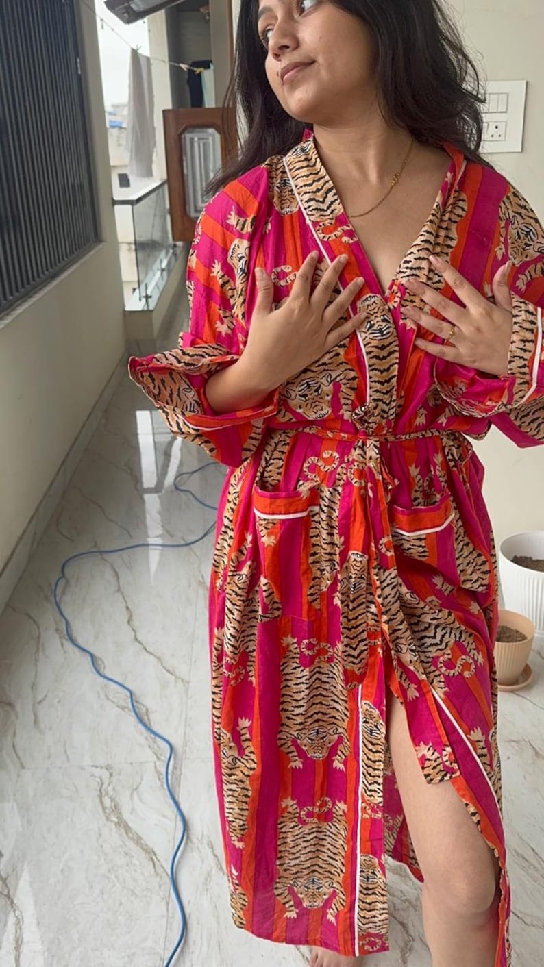 May include: A vibrant, long robe in shades of pink and orange, featuring a tiger stripe pattern. The robe has a wrap-around design with a matching belt, white trim, and pockets. The sleeves are long, and the robe is made of a lightweight fabric.