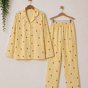 May include: A long-sleeved button-down pajama shirt and matching elastic-waist pajama pants featuring a yellow and white vertical striped pattern with small red heart icons scattered throughout the fabric, displayed on wooden hangers.