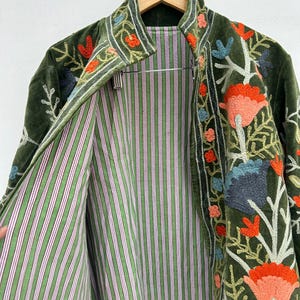 May include: Olive green velvet jacket with floral embroidery in orange, blue, and pink. The jacket's interior features a green and white striped lining. The jacket is hanging on a wooden hanger.