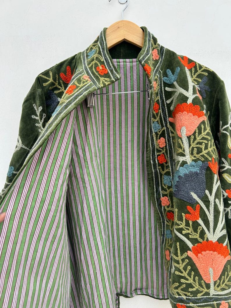 May include: Olive green velvet jacket with floral embroidery in orange, blue, and pink. The jacket's interior features a green and white striped lining. The jacket is hanging on a wooden hanger.