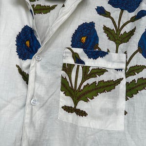 May include: White button-down shirt with a floral pattern. The shirt features large blue flowers with yellow centers and green leaves. A pocket on the chest displays a matching floral design. The shirt is made of a lightweight fabric.